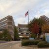 Multiple-story hospital building with trees, parking lot, directional sign, and flag pole with flag waving