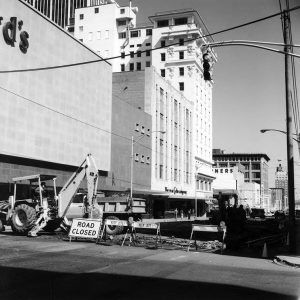 Truck and back hoe loader on city street behind "Road Closed" sign and barriers with multistory buildings on either side