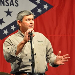 White man speaking at microphone with Arkansas flag background behind him