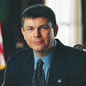 White man in suit and tie with flag behind him