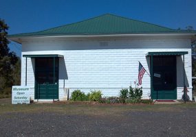 Single-story brick building painted white with green roof and sign in front saying "museum open Saturday"