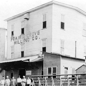 White men standing before large building "Prairie Grove Milling Company"