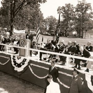 White man on stage speaking at lectern with other white men in suits and flags behind him