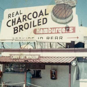 Small brick building with awning with "Real charcoal broiled hamburgers" and "carry out window" signs above it