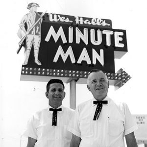 Young white man and older white man in matching white shirt and black pants uniforms with ties under "Wes Hall's Minute Man" sign