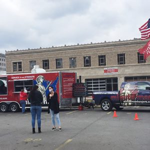 Group of white patrons in parking lot outside red food truck with multistory buildings in the background