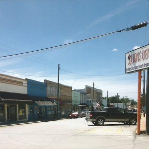 Storefronts on street with parked cars and blue skies and sign reading "Lions Den Roller Skating Food Arcade"