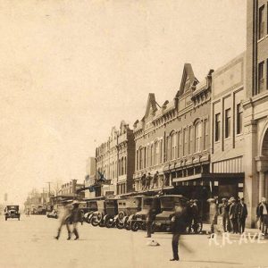 Row of cars parked outside multistory buildings with people crossing the street in the foreground