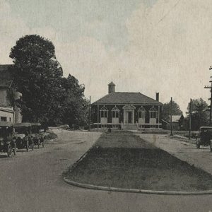 Cars parked next to brick buildings on either side of partition with single-story brick building and water tower in the background