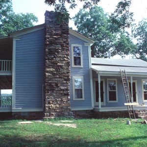 Two story light blue house with covered porch and balcony with brick chimney and a ladder leaning against the porch roof