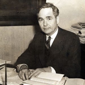 White man in suit and tie with desk covered in papers and open books