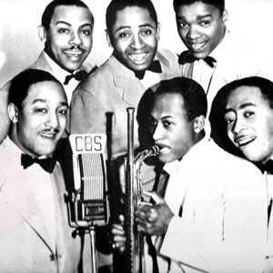 African-American man playing saxophone with five African-American men in suits and bow ties in front of a CBS microphone