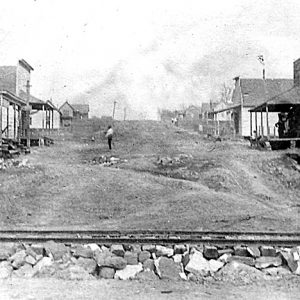 Dirt uphill road with buildings on both sides and man standing and railroad tracks