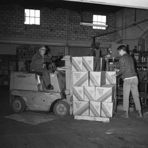 Old white man with glasses and hat moving boxes with forklift in bottling plant