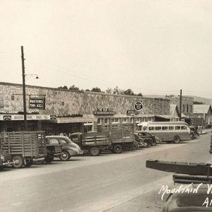 Row of parked vehicles on street outside single-story storefronts with stone walls