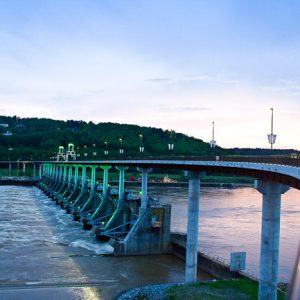 lock and dam on river with pedestrian bridge winding over it and houses, trees on hill in background