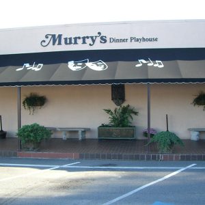 Exterior of "Murry's dinner playhouse" building with decorative plants and benches under its awning