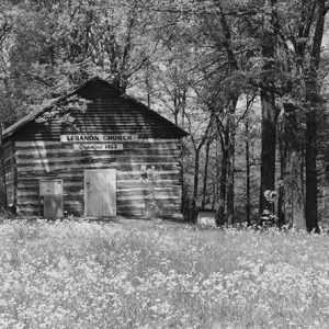 Single-story cabin building with "Lebanon Church organized 1852" sign under trees in field