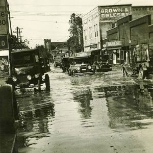 Cars driving on flooded town streets past storefront buildings