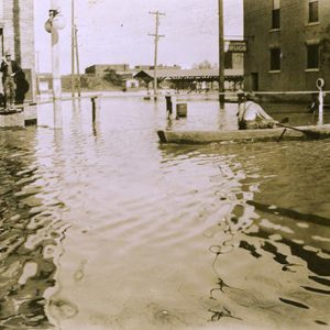 Men standing on boxes while man paddles boat on flooded streets