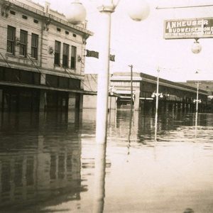 "Anheuser Busch" hanging sign over flooded town street with multistory buildings rising out of the water