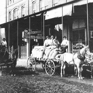 White men with horse drawn wagons on dirt road outside multistory feed and grocery store with covered porch