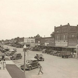 View of town street with parked cars and people walking along sidewalk in front of brick buildings