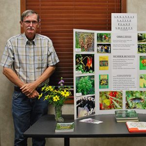 White man with glasses in plaid shirt and jeans standing next to poster display on table