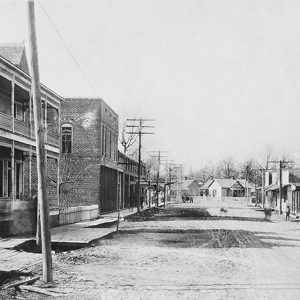 Multistory building with covered porch and balcony on dirt road with houses and storefronts in the background