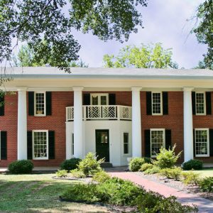 Two-story brick house with covered porch supported by six columns on grass with sidewalk