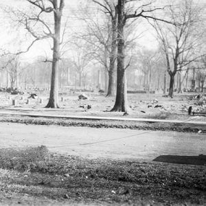 Car parked on dirt road by field with debris under trees in it and people in the background