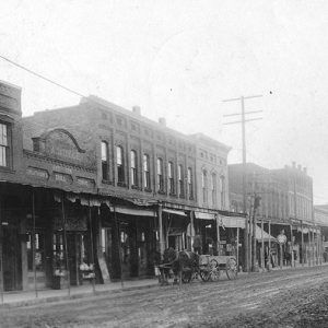 Horse drawn wagon on dirt street outside row of brick storefronts with covered walkways and power lines