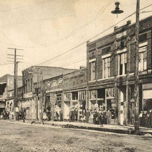 Groups of people standing on sidewalk outside single and multistory buildings on dirt street