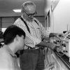 Old white man in suspenders teaching young white man to use lab equipment