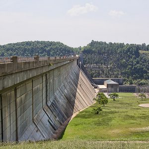 Concrete dam bridge with outbuildings and power station below it with tree-covered mountain in the background