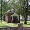 Narrow single-story "Norman Library" brick building with covered porch under trees in park with stone wall and steps in the foreground