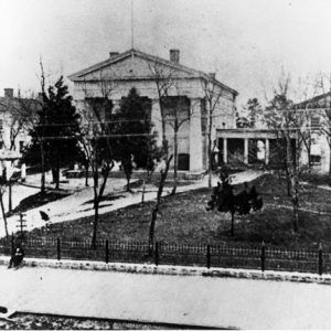 Multistory classical structure with four front-facing column and two smaller wings with road in foreground and seated people on sidewalk