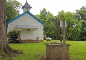 Single-story building with cupola and covered porch on grass with   well in the foreground