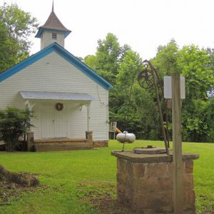 Single-story building with cupola and covered porch on grass with   well in the foreground