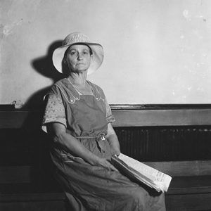 Old white woman sitting in hat and dress with newspaper on her lap