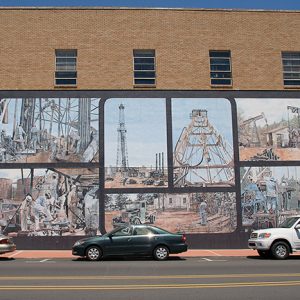 Oil field workers and derricks in mural on side of multistory building on street with parked cars