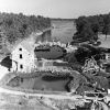 Stone mill building with bridge over water and walking paths and wooden fencing with trees in the background