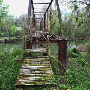 View looking across abandoned steel truss bridge over river with broken overgrown wooden landing in the foreground