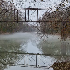 Steel truss bridge reflected in foggy river with trees on both sides