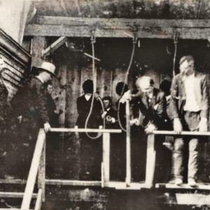 White men in suits inspecting nooses at gallows with hooded men waiting behind them