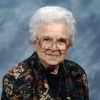 Old white woman with glasses in multicolored jacket and heart shaped pendent necklace with blue background