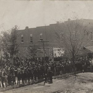 Large crowd gathered on town street and empty lot adjacent to two-story storefront