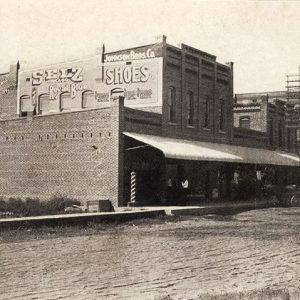 Horse drawn wagons on dirt road outside brick buildings with covered walkway