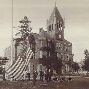 Group of white men taking down large American flag on grounds of multistory building with bell tower