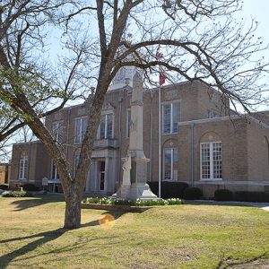 Multistory building with clock tower and stone monument with flag pole and trees in its front yard